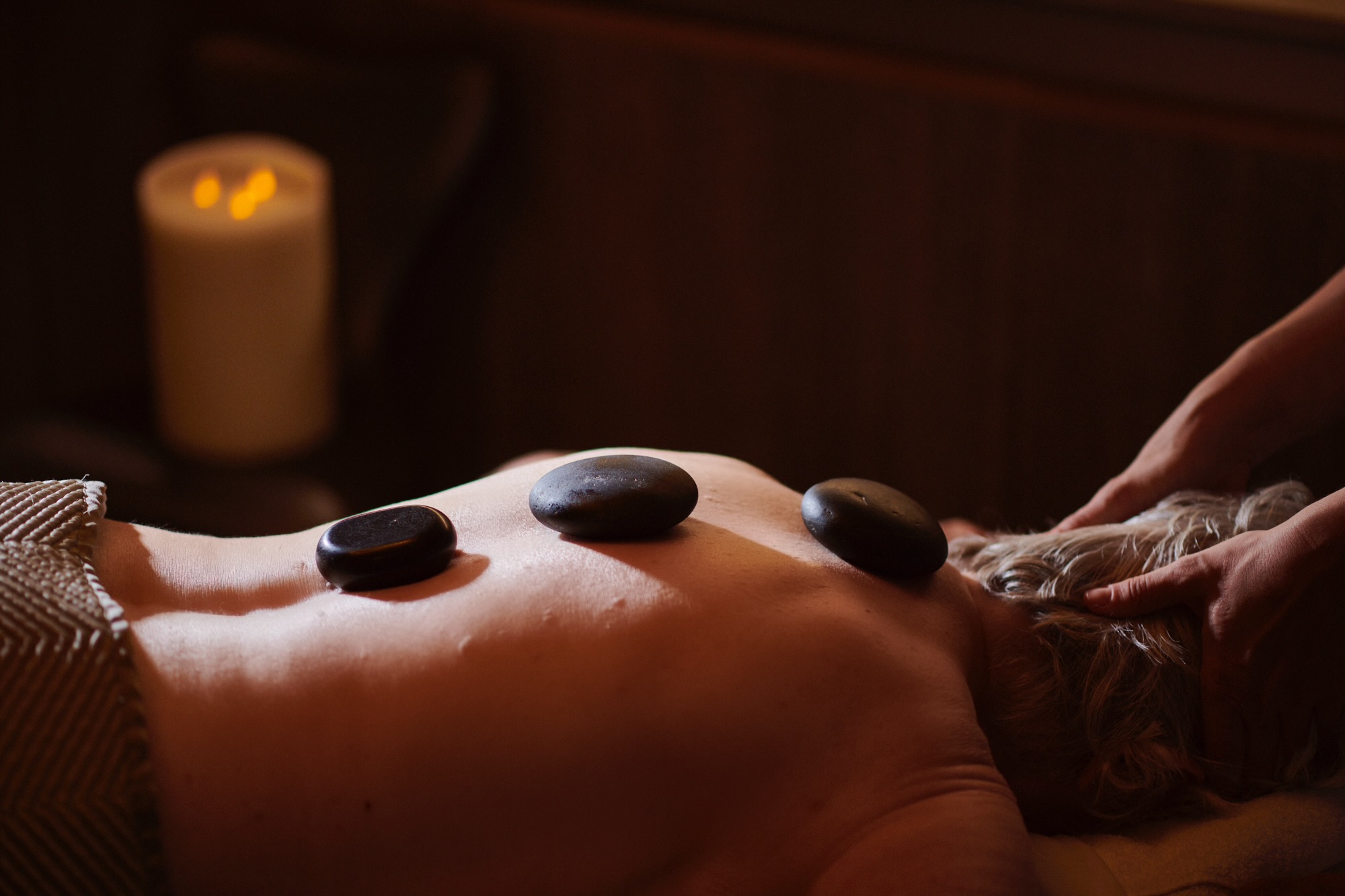 A person is lying face down on a massage table with three stones placed along the spine.