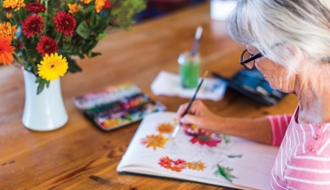 GREY HAIRED WOMAN IN PINK TOP AND GLASSES PAINTING AT WOODEN TABLE, BRIGHTLY COLOURED FLOWERS IN THE BACKGROUND