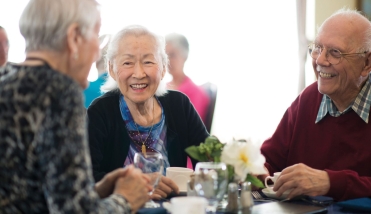 THREE ELDERLY PEOPLE SMILING SITTING AT TABLE ENJOYING DINNER 